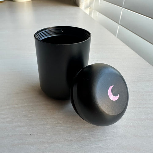 Matte black aluminium capsule jar with its threaded lid tipped open, blush moon on the cap.