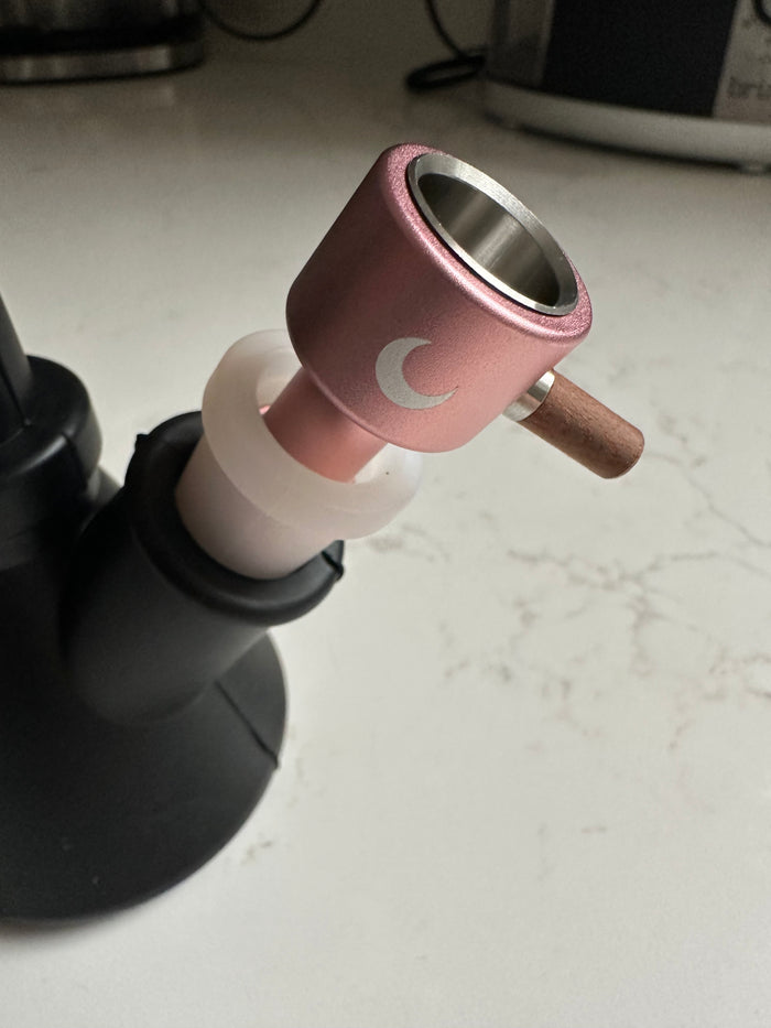 Rose-gold bowl with a wooden side grip and white moon mark, seated in a matte black piece on a marble counter.