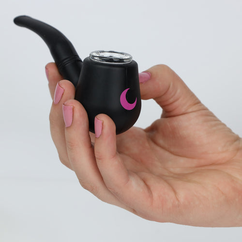 Matte black silicone tobacco-style pipe with a clear glass bowl and blush moon mark, held in a manicured hand against soft white.