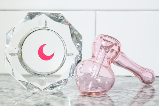 A pink glass hammer bubbler resting beside a faceted crystal ashtray with a pink crescent moon, on a marble surface