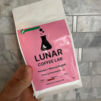 Lunar Coffee Lab Yemen Mocca Hajjah in a white pouch with a bright pink label and the moon-and-flask mark, held against marble tile.