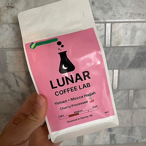 Lunar Coffee Lab Yemen Mocca Hajjah in a white pouch with a bright pink label and the moon-and-flask mark, held against marble tile.