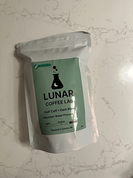 Lunar Coffee Lab Half Caff dark blend in a white pouch with a soft green label and the moon-and-flask mark, on white marble.