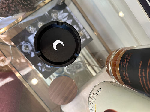 Round matte black metal ashtray with a white moon mark, styled on a mirrored vanity tray beside perfume bottles.