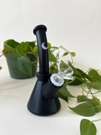 Matte black silicone vase-shaped water piece with a blush moon mark and clear glass bowl, framed by trailing pothos leaves.