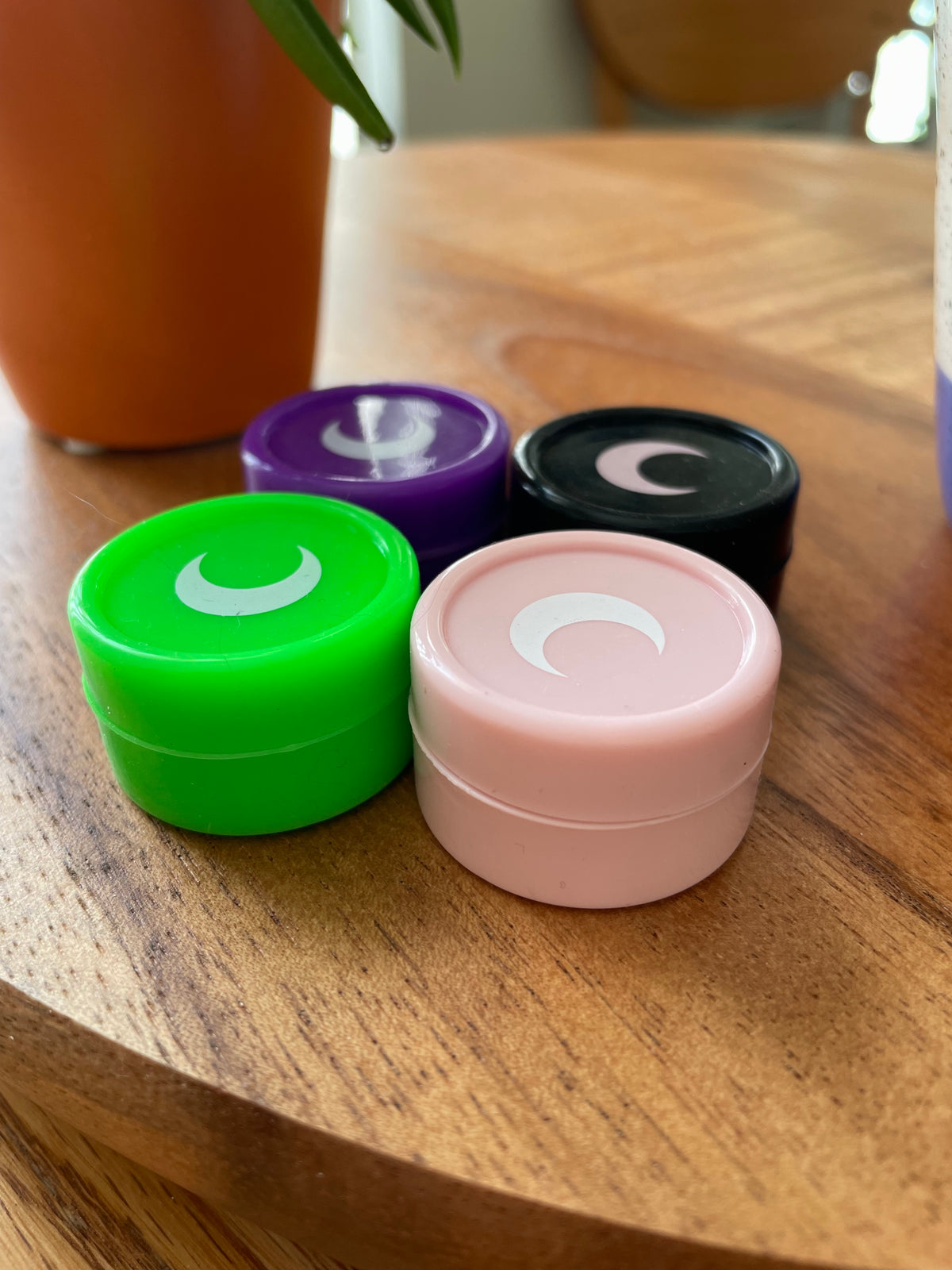 Four small silicone jars in green, purple, black and blush, each capped with a white moon, on a wood table.