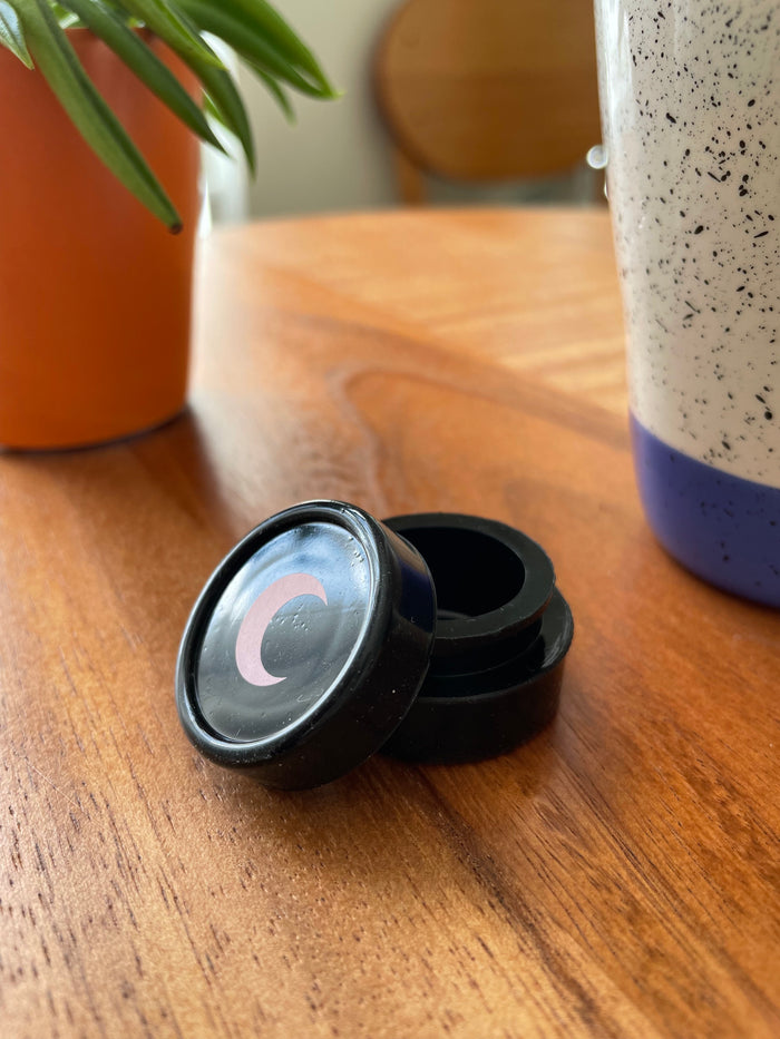 Small black silicone jar with its lid tipped open, blush moon on the cap, on a warm wood table.