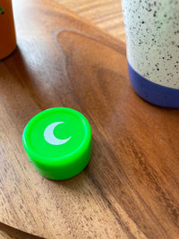 Small green silicone jar with a white moon on the lid, on a wood table beside a speckled vase.