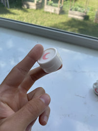 Small white silicone jar with a blush moon on the lid, held between finger and thumb in window daylight.