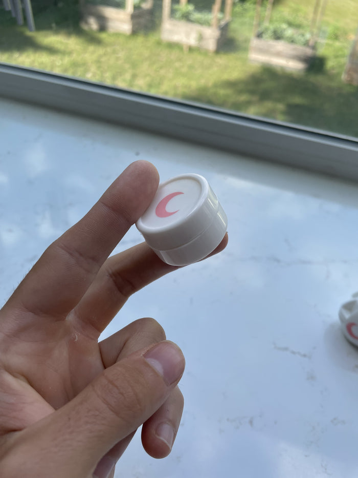 Small white silicone jar with a blush moon on the lid, held between finger and thumb in window daylight.