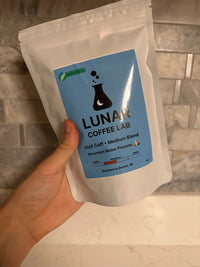 Lunar Coffee Lab Half Caff medium blend in a white pouch with a sky-blue label and the moon-and-flask mark, held against a marble counter.