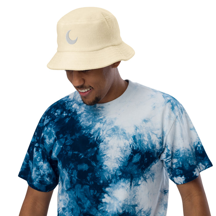 Cream terry cloth bucket hat with a white crescent moon embroidered at the front, worn with a blue tie-dye tee.
